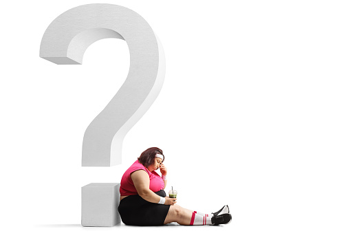 Sad overweight woman in sportswear holding a green shake and sitting against a big question mark isolated on white background