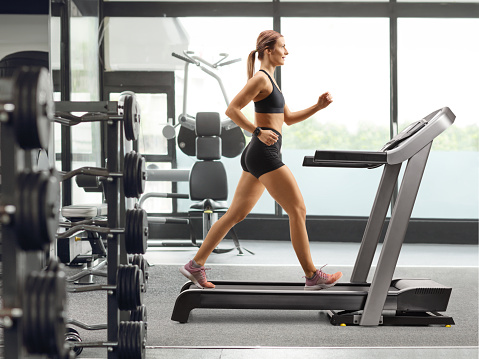 Full length profile shot of a muscular young woman walking on a treadmill at a gym