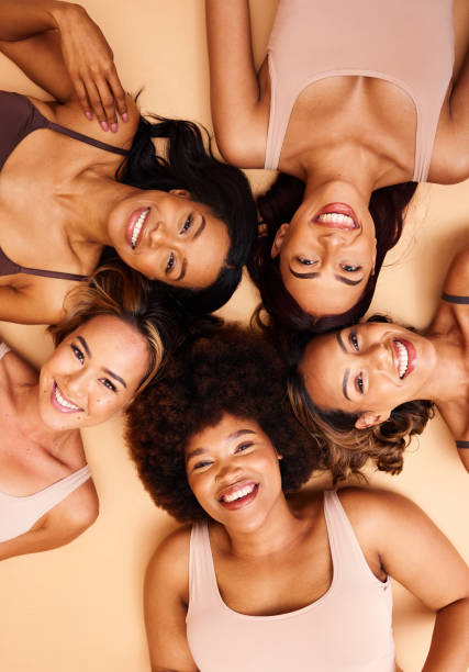 Beauty, above and portrait of a diversity of women with smile for skincare isolated in studio brown background together. Support, happy and group of friends on a floor for skin, care and face makeup Beauty, above and portrait of a diversity of women with smile for skincare isolated in a studio brown background together. Support, happy and group of friends with care laying on floor for skin diverse models stock pictures, royalty-free photos & images