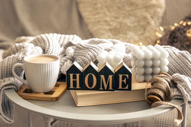 Cozy composition with candles, coffee cup, book and decorative word home on blurred interior background.
