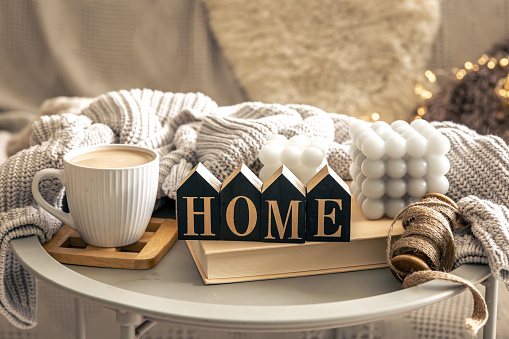 Cozy composition with candles, coffee cup, book and decorative word home on blurred interior background.