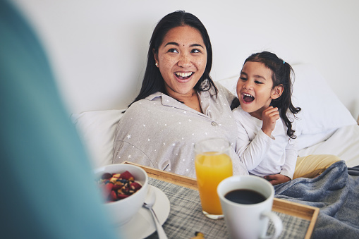 Mother and child with breakfast in bed in the bedroom for mothers day surprise at home. Happy, smile and young mom relaxing with girl kid with a healthy meal for brunch on a weekend at their house.