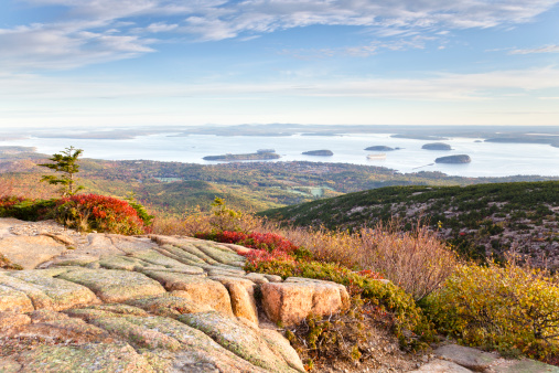 An early morning view of Frenchman Bay from the top of Cadillac Mountain in Autumn; Acadia National Park, Maine. Cadillac Mountain's granite rocks and autumn-colored red ground cover are in the foreground, while the background has the small town of Bar Harbor, along with the Porcupine Islands and Bar Island in Frenchman Bay.
