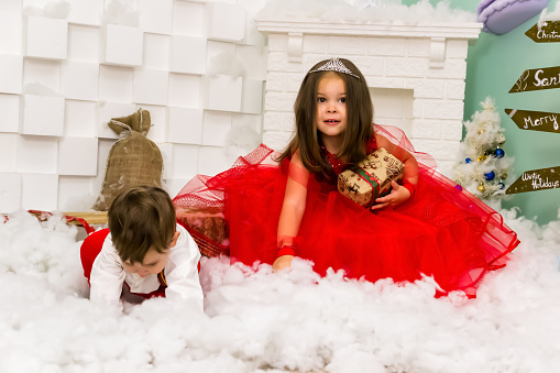 A little girl and a boy in red clothes have fun in the artificial snow by the fireplace in the studio. The girl is holding a box with gifts. Little kids in red christmas costume having fun beside a decorated christmas tree. Two kids playing with falling artificial snow flakes near a christmas tree.Kids playing with artificial snow flakes falling near a christmas tree. Christmas concept. High quality photo.