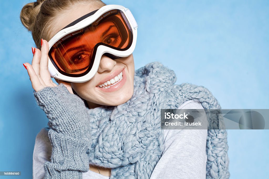 Winter Girl Winter portrait of a beautiful young woman wearing goggles and scarf posing against blue background. Looking at camera and smiling. Studio shot. Fashion Model Stock Photo