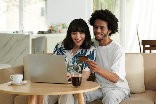 Happy young intercultural couple choosing online goods in front of laptop while African American man with credit card going to pay for purchase