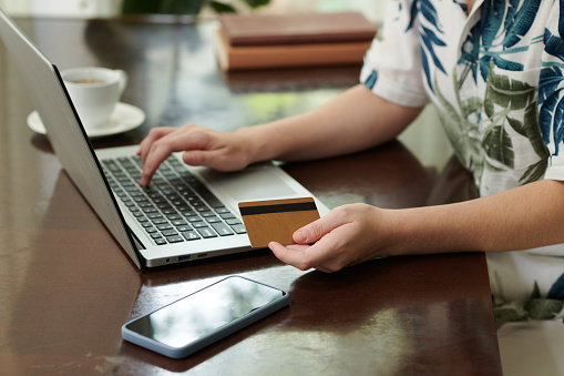 Focus on hand of young female shopper with credit card sitting by table in front of laptop and entering banking data during contactless payment