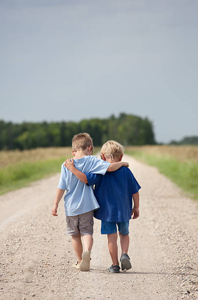 Two Boys Walking Down a Gravel Road Two young boys with their arms around each other walking down the country road. Vertical colour image. Copyspace. Back view of two cute Caucasian boys. Additional themes include friendship, bonding, talking, care, love, siblings, brothers, walking, rural, exercising, togetherness, cute, humour, summer, relationships, and affection. blue brothers stock pictures, royalty-free photos & images
