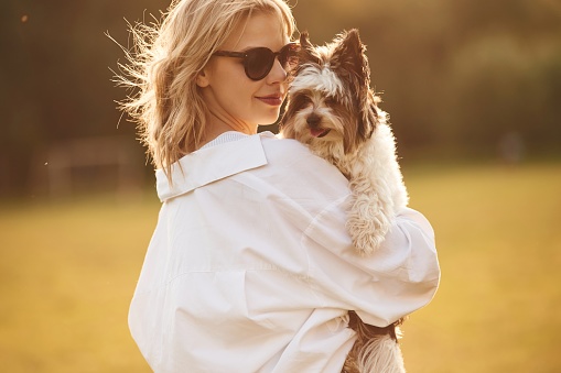 Woman in sunglasses is standing on the summer field and holding little dog.