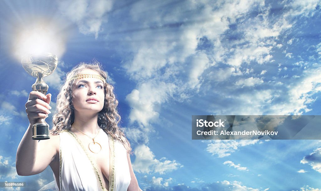 The Greek Goddess Carrying a Goblet of Fire. Toned Portrait of The Beautiful Young Woman Holding a Gold Goblet of Flame in the Blue Sky. She is Wearing the White Greek Tunic. Achievement Stock Photo