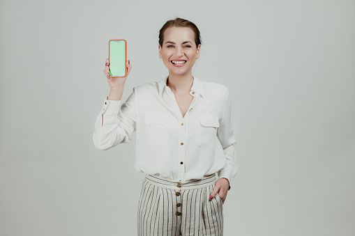Portrait of a smiling woman wearing a white shirt holding a smartphone with a green screen, studio shot in front of a white background