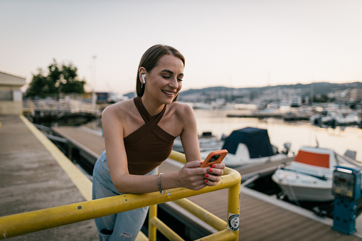 Happy and cheerful young woman using a smartphone and in-ear headphones at the seaport on a lovely summer day in Kavalla