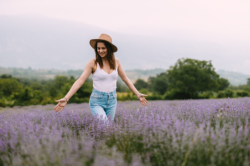 A beautiful young woman wearing casual clothes and a hat walking through the lavender field on a lovely summer day