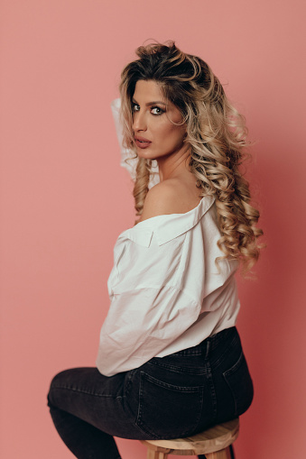 Portrait of a beautiful young fashionable woman with long blond hair sitting on a wooden stool, studio shot in front of pink background