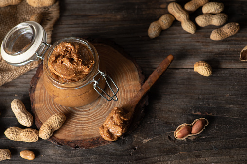 Freshly made homemade peanut butter in a glass jar and raw peanuts in a wooden scoop, selective focus o dark vooden background