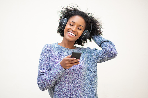 Portrait of happy young black woman listening to music with mobile phone and headphones against white background