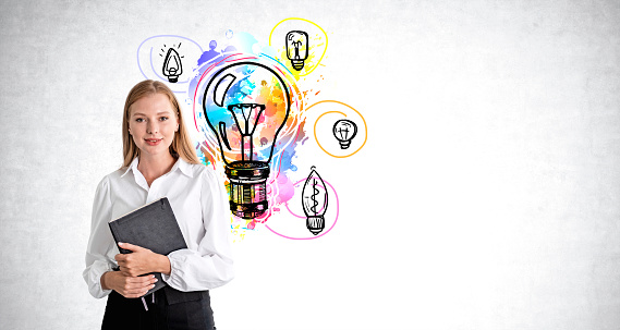 Happy young businesswoman with notebook in hands, different colorful light bulbs doodle on grey concrete wall background. Concept of business startup and idea