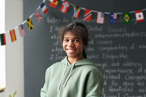 Cheerful Hispanic private middle school student smiles before class. She is standing in her classroom. He has a braid and is wearing a backpack and a school uniform. Students are in the background talking.