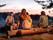 A company of young people sit around a fire at night on the banks of the river and relax in nature. Cooking food on a fire, outdoors