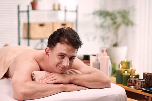 Happy young multiethnic man looking at camera while relaxing on massage couch or table and keeping chin on rolled soft towel