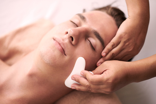 Young relaxed male client of spa salon getting luxurious gua sha facial massage bwing given to him by professional female masseuse