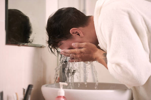 istock Young man in soft white bathrobe bending over sink and washing his face 1659063388