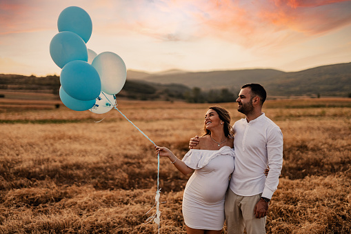 A beautiful pregnant woman and a young man dressed in white holding gender-reveal balloons in nature on a sunny summer day