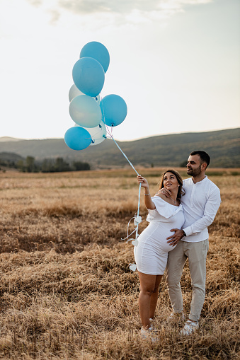 A beautiful pregnant woman and a young man dressed in white holding gender-reveal balloons in nature on a sunny summer day