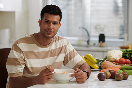 Happy young Indian man in casualwear looking at camera while sitting by table in the kitchen and having muesli from bowl during breakfast
