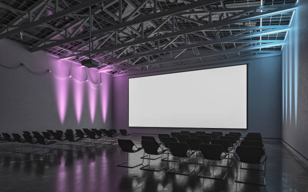 istock Side view of an empty darkened large gallery/showroom interior with empty seats (no people), and a wide projection screen in the middle for events under a ceiling industrial construction, illuminated by multi-colored side wall reflectors 1659130251