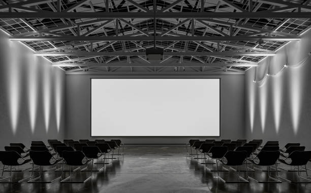 istock Front view of an empty darkened large gallery/showroom interior with empty seats (no people), and a wide projection screen in the middle for events under a ceiling industrial construction, illuminated by side wall reflectors 1659130277