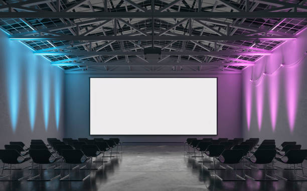 istock Front view of an empty darkened large gallery/showroom interior with empty seats (no people), and a wide projection screen in the middle for events under a ceiling industrial construction, illuminated by multi-colored side wall reflectors 1659130297
