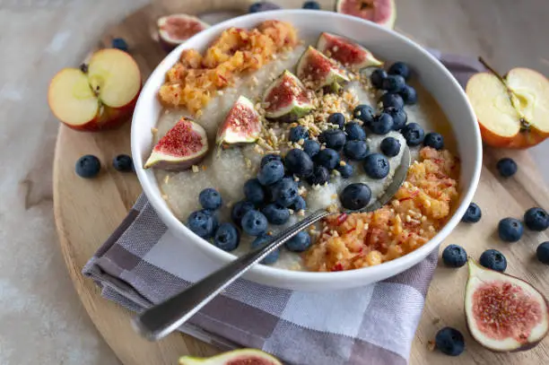 Homemade breakfast bowl with fresh cooked millet porridge. Cooked with water for vegan breakfast. Top with fresh fruits and honey. Served in a bowl with spoon on rustic background with decoration. closeup