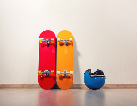 Two skateboards, yellow and red, are standing against the wall, a blue protective helmet is lying next to it, the floor is wooden.