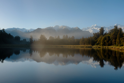 Pre-dawn mist drifts across the mirror-like surface of spectacular Lake Matheson on New Zealand’s South Island. whilst beyond we see the snow capped peaks of Mt Tasman and Mt Cook and the Southern Alps.