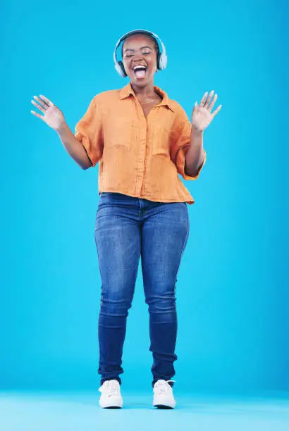 Photo of Black woman, headphones and singing to music with dance in a studio. Audio streaming, radio and happy female person with blue background and excited from celebration and freedom with hip hop song