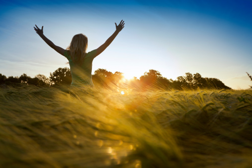 Woman holding arms up to the setting sun while standing in a field of barley.