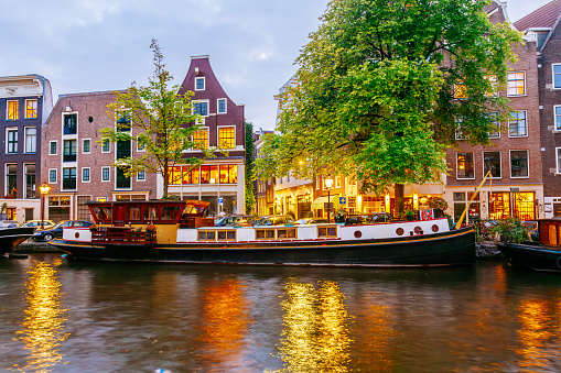A canal in Amsterdam at dusk. The canal is lined with trees and buildings on both sides. The buildings are traditional Dutch style with gabled roofs and large windows. Several boats are moored along the canal. The water is calm and reflects the lights from the buildings and boats. The sky is a deep blue with a few clouds.