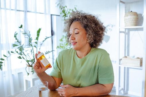 Shot of mature woman in green t-shirt sitting at the table in the living room and holding bottle od pills in her hand