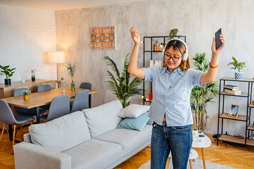 Cheerful Asian woman listening to music on headphones and dancing at home.