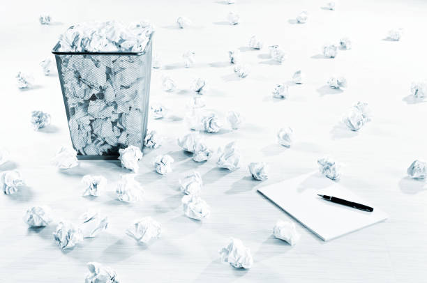 Many crumpled paper balls scattered on the white floor, overflow paper bin, blank notepad and pen.