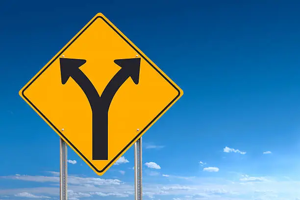 An informational traffic sign over a blue sky showing a division of directions - choice or decision - a clipping path is included to separate sign from bkg. Sign positioned to the left of the frame allowing for plenty of copy space.