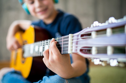Close-up of cute small boy whit headphones learning to play guitar while sitting at home. Focus is on hand