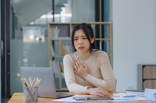 An Asian female has a heart attack pain, and tension-sick employee who is feeling heartache while working on a laptop at the workplace at a desk in the office.