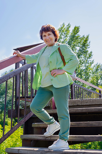 Attractive smiling elderly woman stands on the bridge on a summer sunny day against the backdrop of nature