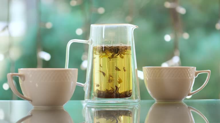 Teapot with herbal tea and two white cups on the table outdoors. Pouring green hot tea into white cups