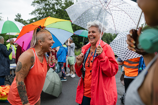 A medium close up front view of a group of women who are stood waiting for a park run to start. They are using colourful umbrellas to keep themselves dry and are enjoying a catch up before the warm up exercise starts.