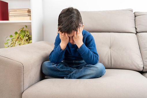Upset problem child with head in hands sitting on sofa concept for childhood bullying, depression stress or frustration
