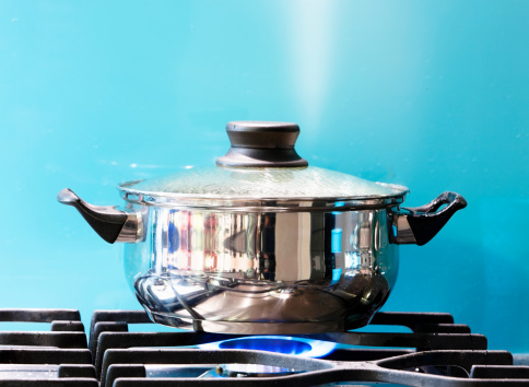 A covered pot on a lit gas burner shows a jet of steam rising as it boils against a turquoise wall.