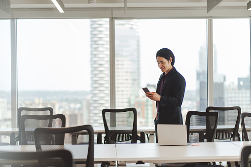 Portrait of smiling Asian businessman wearing stylish black suit holding mobile phone standing in meeting room. Japanese male checking mail, browsing. Technology concept